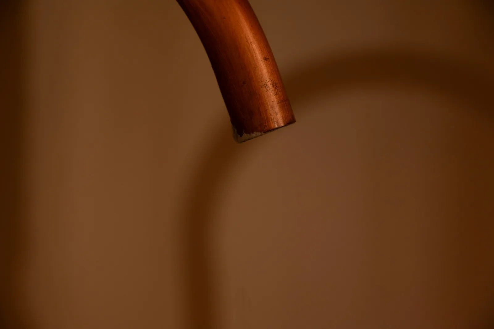 One copper pipe curves down in front of a brown wall and stops at a cut end.