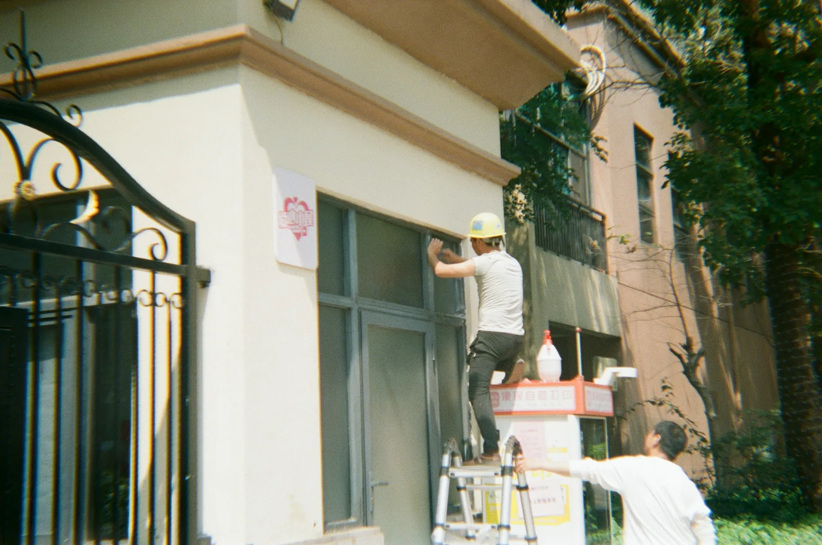 One man in a hard hat works at a window frame up a ladder, another steadies it.