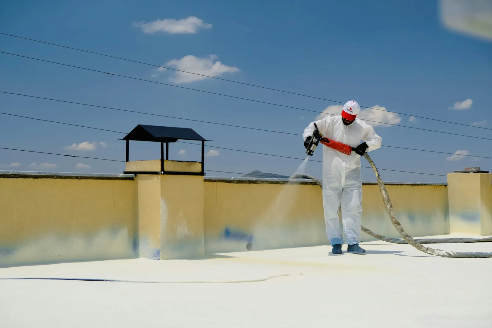 Spray gun in hand, hose trailing behind him, a man in coveralls coats a flat roof white.