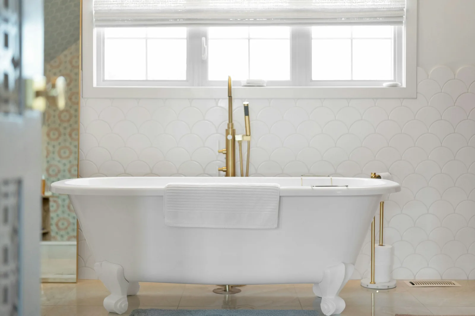 A freestanding white bath beneath a window in a tiled bathroom.