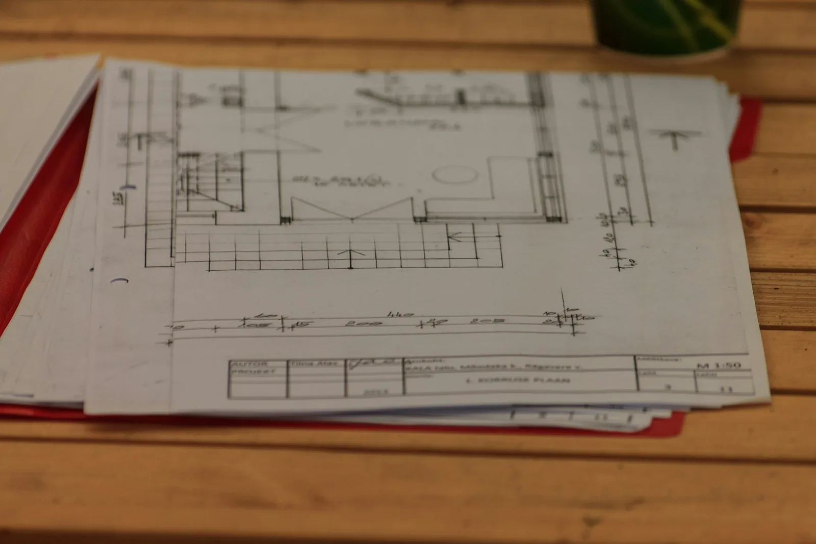 A pencil floor plan at 1:50 lies open in a red folder on a slatted timber table.