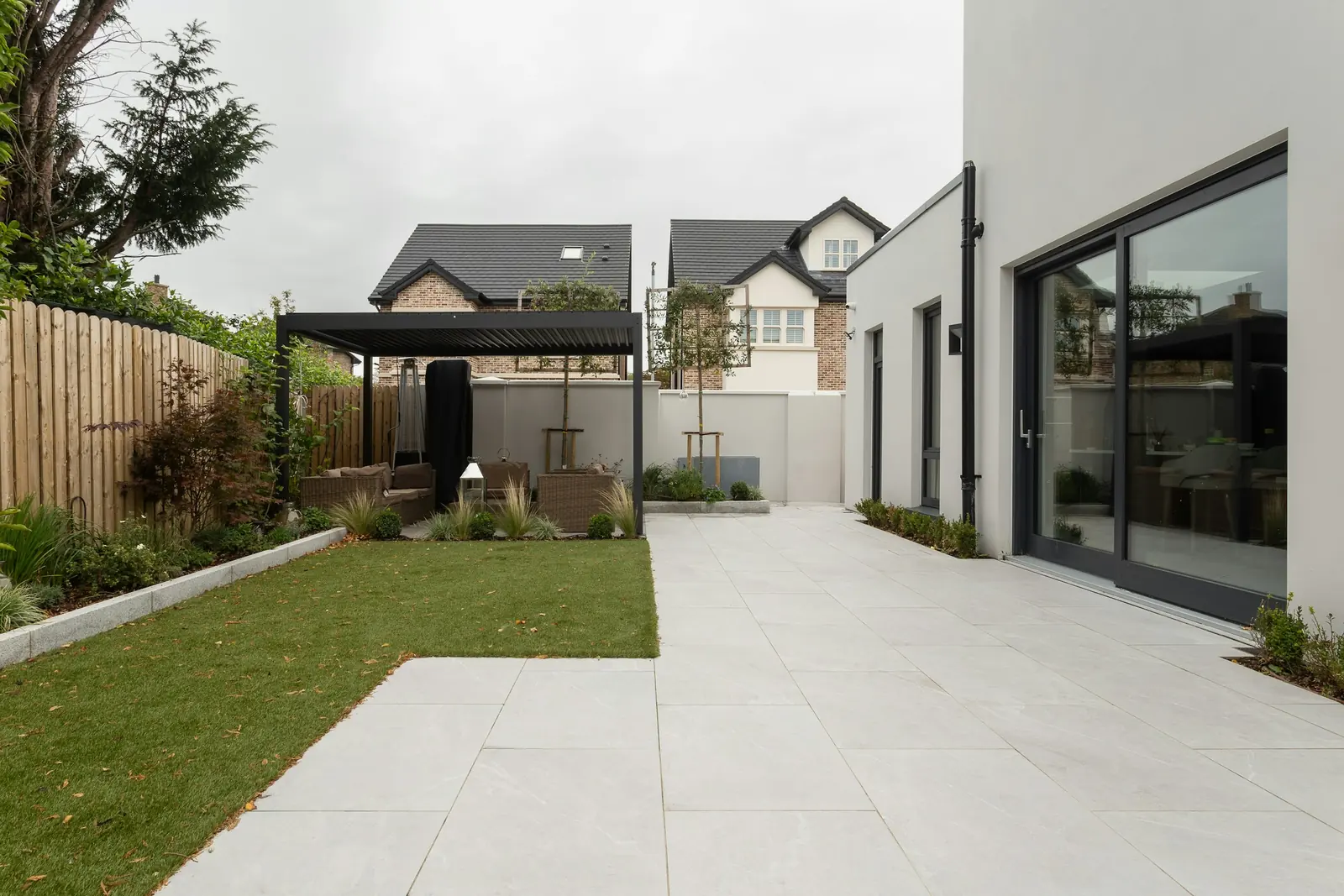 Rattan seating sits under a black louvred pergola at the far end of the paved garden.
