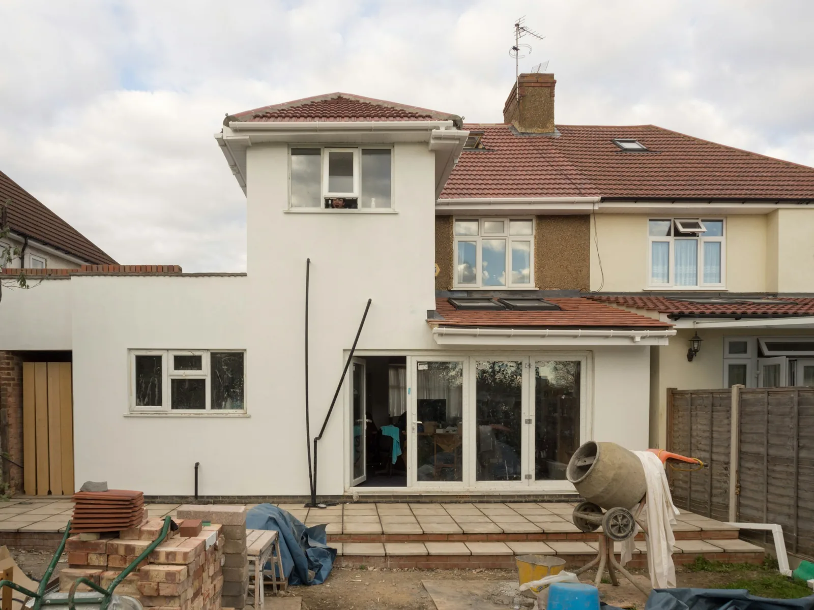 Someone looks out of the new upstairs window; roof tiles and a mixer still sit in the garden.