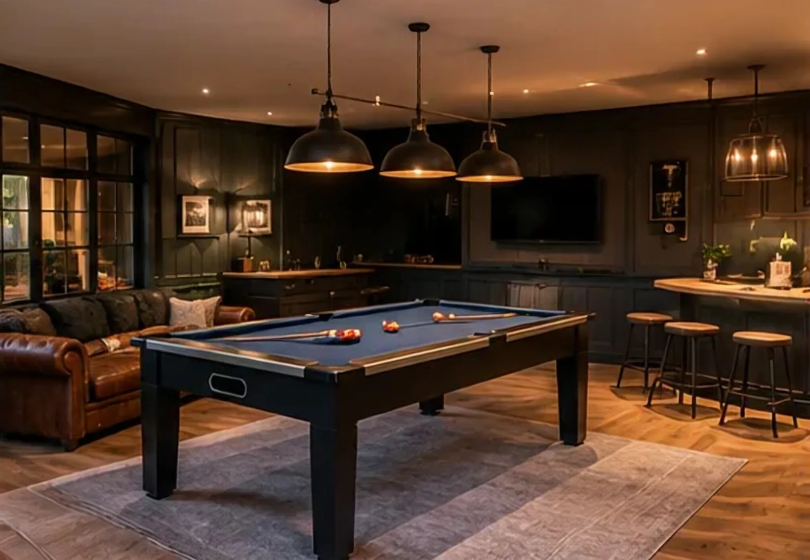Inside a garden room: a pool table under three pendant lamps, a leather sofa, and a small bar with stools against dark panelled walls.