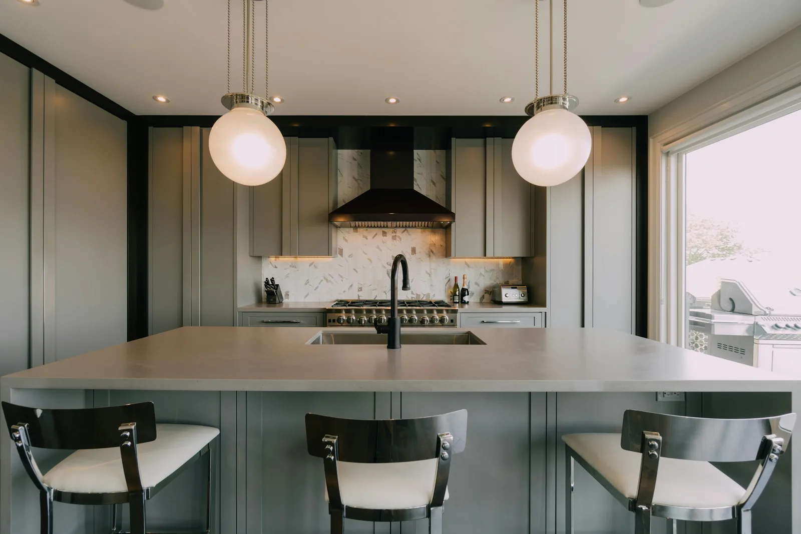 A dark green kitchen with a central island, pendant lights and bar stools.
