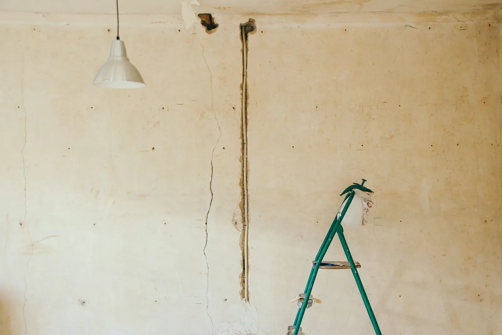 A cracked plaster wall in a room being refurbished, with a stepladder and a bare pendant light.
